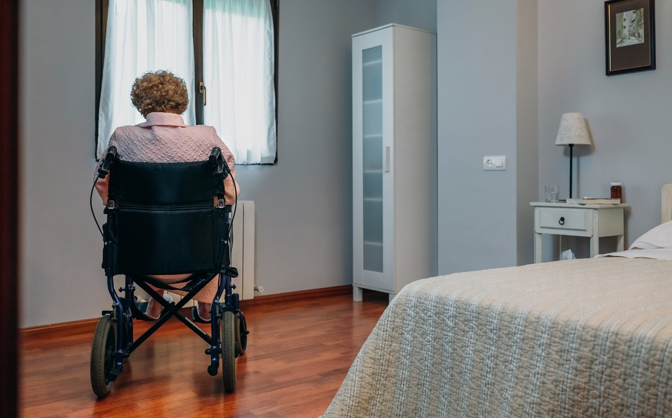 An older woman in a wheelchair sits alone in a nursing home room, facing a window with sheer curtains. A bed, nightstand, lamp and cabinet are visible in the quiet room.