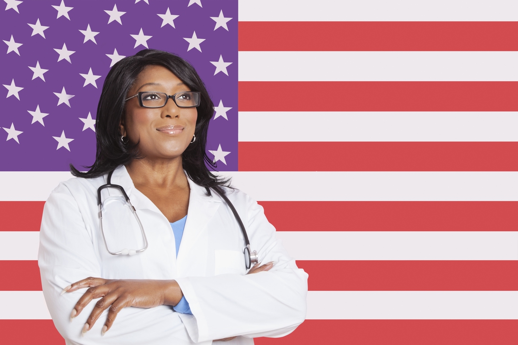 A Black woman physician wearing glasses, a white medical coat, and a stethoscope stands with her arms crossed in front of a stylized American flag with purple stars and red stripes. The image represents healthcare freedom and the call for universal Medicare for All discussed in this week’s Code WACK! podcast.