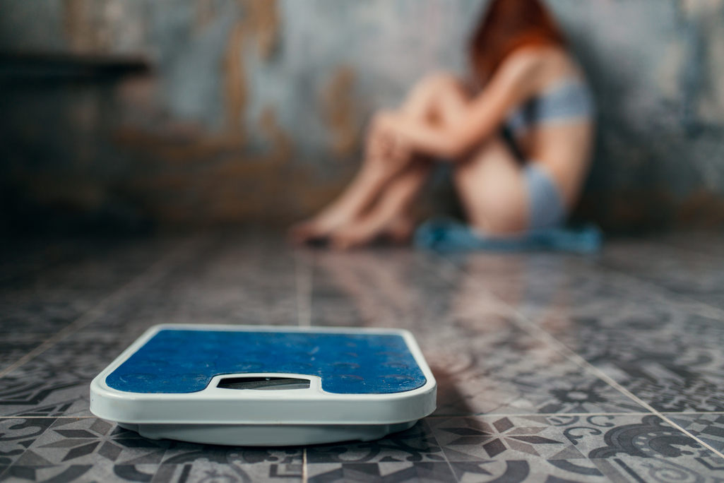 Blue bathroom scale in the foreground with a blurred seated person in the background.