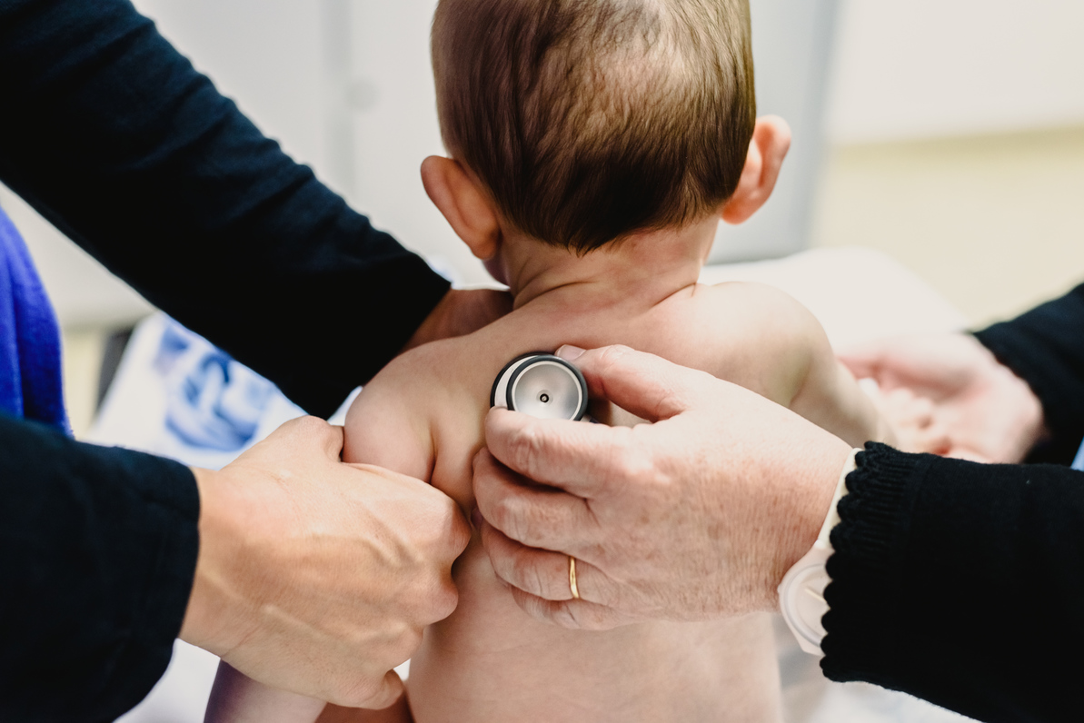 Healthcare professional uses a stethoscope to examine an infant’s back while another adult supports the child.
