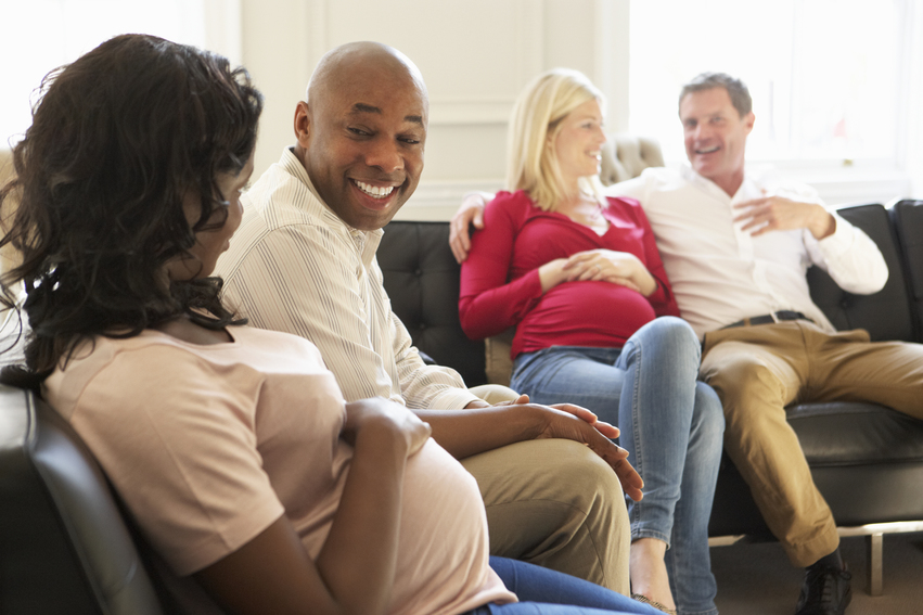 Four adults, including two pregnant women, sit together talking in a bright room.