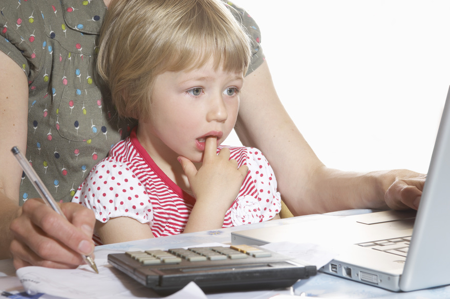 Young child beside an adult working with a laptop, calculator, paperwork, and pen at a table.