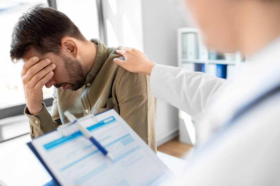 A distressed man covers his face while a healthcare professional rests a hand on his shoulder, with medical paperwork in the foreground.