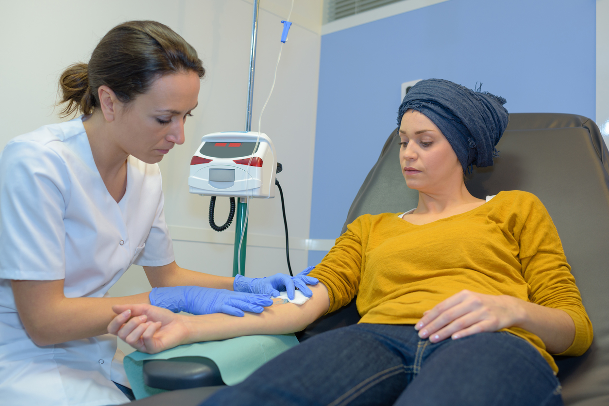 Healthcare worker attends to tubing at a seated person’s arm beside an IV pole in a clinical room.