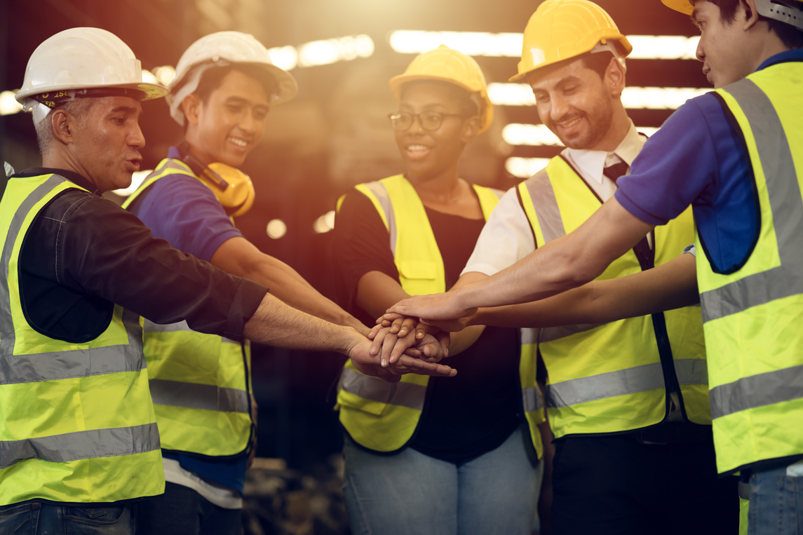 A group of workers wearing hard hats and high-visibility safety vests stand together with their hands stacked in the center. The workplace setting and gesture of solidarity connect visually with this week’s discussion of labor, collective bargaining, and employer-sponsored healthcare.
